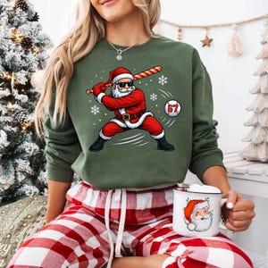 May include: A green sweatshirt featuring a graphic of Santa Claus in a baseball stance, swinging a bat. Santa is wearing sunglasses and a red suit. A baseball with the number 67 is in the image. The person is wearing red and white plaid pajama pants and holding a Santa mug.