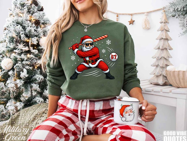 May include: A green sweatshirt featuring a graphic of Santa Claus in a baseball stance, swinging a bat. Santa is wearing sunglasses and a red suit. A baseball with the number 67 is in the image. The person is wearing red and white plaid pajama pants and holding a Santa mug.