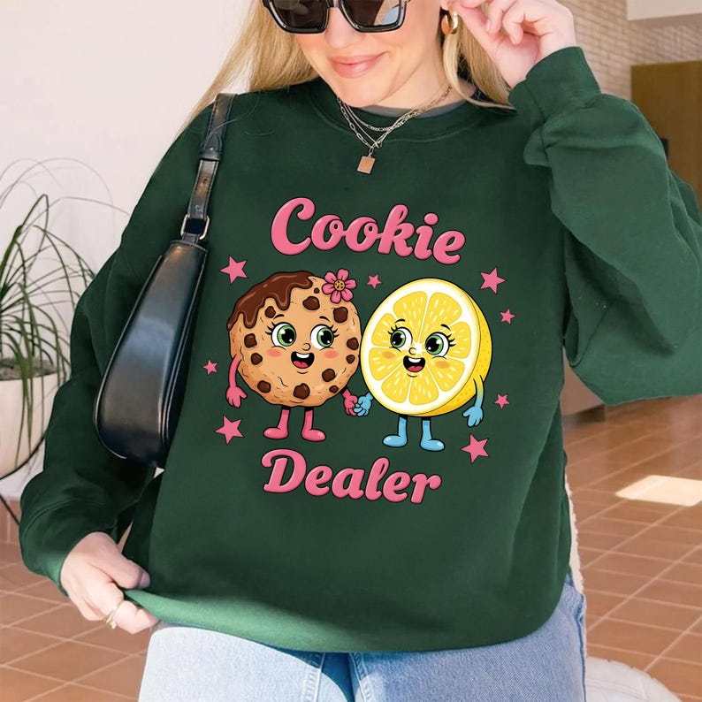 Puede incluir: Sudadera verde oscuro con el texto "Cookie Dealer" en rosa. El gr&aacute;fico presenta una galleta y un lim&oacute;n sonrientes tomados de la mano, rodeados de estrellas rosas. La galleta tiene glaseado de chocolate y una flor.