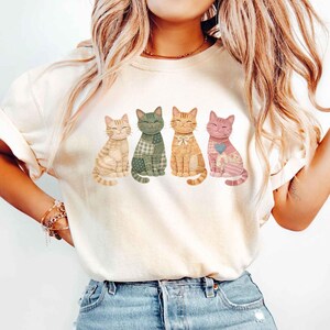 May include: Cream-colored t-shirt featuring four smiling cat illustrations. Each cat has a unique design, including stripes, plaid, and floral patterns, in shades of beige, green, orange, and pink. The cats are sitting upright, with their tails curled.