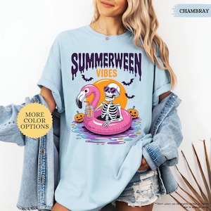 K&ouml;nnte beinhalten: Hellblaues T-Shirt mit dem Text "SUMMERWEEN VIBES" und einer Grafik eines Skeletts mit Sonnenbrille, das auf einem rosa Flamingo-Schwimmring schwimmt. Das Design zeigt Flederm&auml;use, K&uuml;rbisse und eine Sonne.