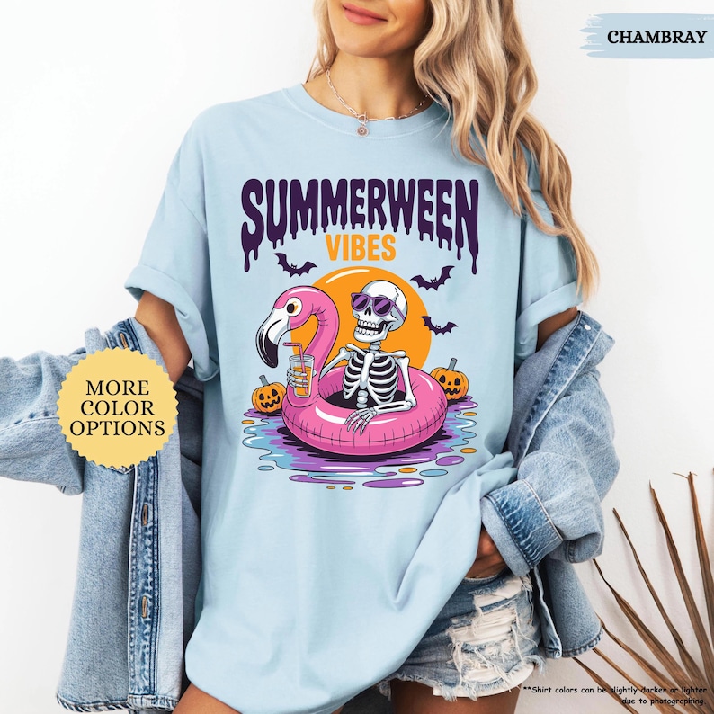 K&ouml;nnte beinhalten: Hellblaues T-Shirt mit dem Text "SUMMERWEEN VIBES" und einer Grafik eines Skeletts mit Sonnenbrille, das auf einem rosa Flamingo-Schwimmring schwimmt. Das Design zeigt Flederm&auml;use, K&uuml;rbisse und eine Sonne.