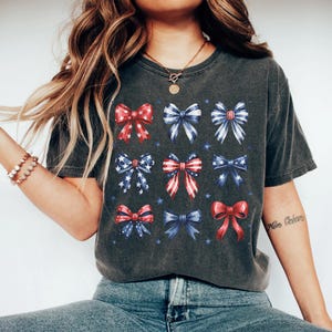May include: A dark gray t-shirt with a pattern of red, white, and blue bows. The bows are arranged in a grid pattern and have a vintage look. The shirt has a relaxed fit and is perfect for a casual summer outfit.