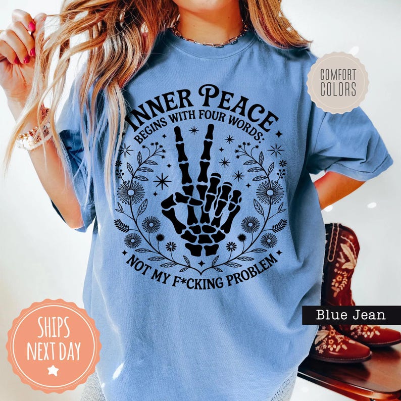 Puede incluir: Camiseta azul claro con el texto "INNER PEACE BEGINS WITH FOUR WORDS: NOT MY F*CKING PROBLEM" y un gr&aacute;fico de mano de esqueleto negro. La camiseta tiene cuello redondo y mangas cortas.