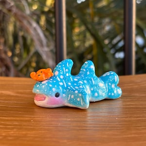 Può includere: Una statuetta di squalo balena blu e bianca con un piccolo pesce arancione sulla testa. La statuetta è fatta di argilla e ha una finitura dipinta a mano.