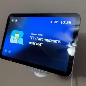 Puede incluir: Un dispositivo doméstico inteligente con pantalla muestra una interfaz azul con el texto "Find art museums near me". La pantalla también muestra la temperatura a 30 grados y la hora 10:59. El dispositivo está montado en una pared.