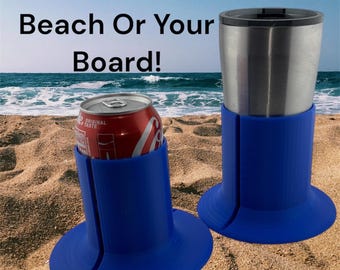 May include: Two blue silicone drink holders with a can and a tumbler on a sandy beach. The holders have a wide base and a narrow top to hold the drinks securely. The text "Great On The Beach Or Your Board!" is visible at the top of the image.