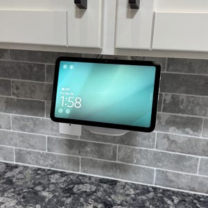 May include: A smart home device with a screen displaying the time and temperature, mounted on a gray brick wall. The device is white and black, with a light blue screen. The time is 1:58 PM.