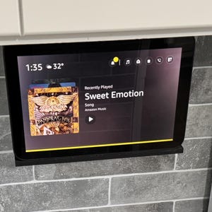 May include: A black smart display mounted on a gray tiled wall, displaying the time, temperature, and music information. The screen shows the song "Sweet Emotion" and album art. A white cable and a white stove are visible.
