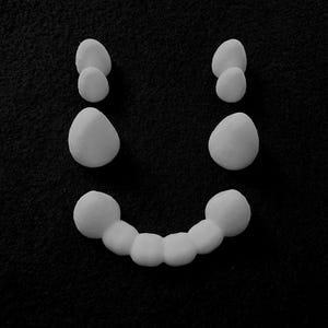 May include: A monochrome image showing a smiley face constructed from white, rounded forms. Two pairs of smaller shapes create the eyes, with a larger shape beneath each, and a curved line of connected shapes for the mouth, set against a black backdrop.