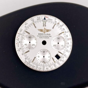 Könnte beinhalten: Ein silbernes Breitling Chronometre Navitimer Zifferblatt mit mehreren Hilfszifferblättern und Markierungen. Das Zifferblatt zeigt das Breitling-Logo sowie Text, der Geschwindigkeits- und Entfernungsmaße in MPH und KM angibt. Das Zifferblatt befindet sich vor einem schwarzen Hintergrund.