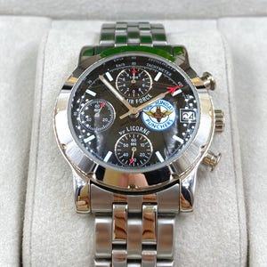 Op de afbeelding: Een roestvrijstalen polshorloge met een zwarte wijzerplaat met meerdere sub-wijzerplaten en de tekst "AIR FORCE" en "SUNDAY PUNCHERS". Het horloge heeft een zilveren band en een datumweergave op 3 uur.
