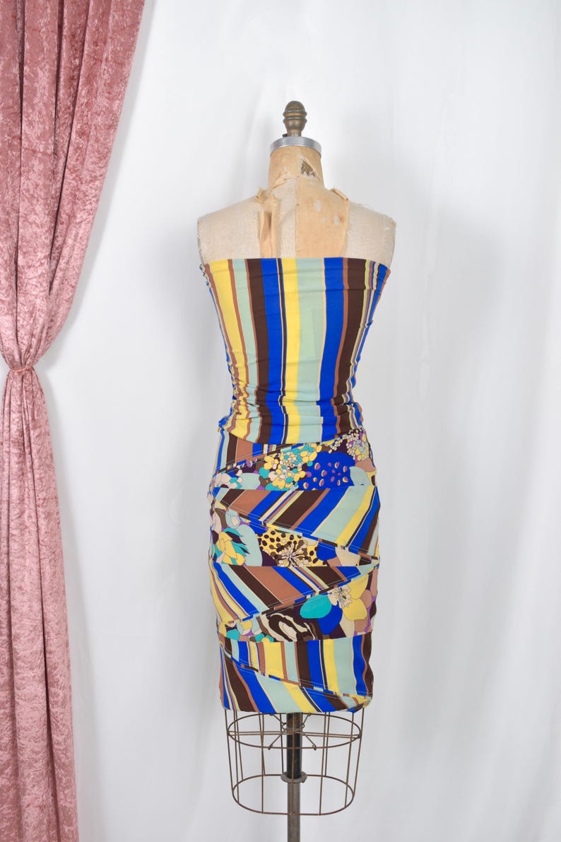 Puede incluir: Vestido sin tirantes y colorido sobre un maniqu&iacute;. El vestido presenta rayas verticales en azul, amarillo y marr&oacute;n en el corpi&ntilde;o, que se transforman en una falda estampada con dise&ntilde;os florales y geom&eacute;tricos. El vestido es fruncido y ajustado.