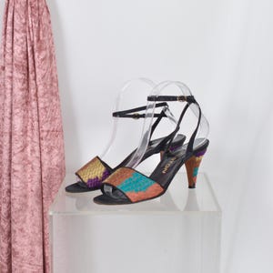 May include: Black high-heeled sandals with patterned uppers. The open-toe shoes have ankle straps and a heel height of approximately 7.6 cm. The uppers feature gold, purple, teal, and orange patterns. The heels are also patterned.