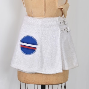 Puede incluir: Minifalda de rizo blanca con un parche circular con rayas azules, blancas y rojas. La falda tiene detalles de hebillas plateadas en el lateral y se muestra en un maniquí.