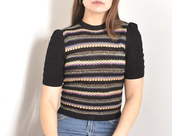May include: A woman wearing a black and multi-colored striped short-sleeved sweater with a round neckline and blue jeans.