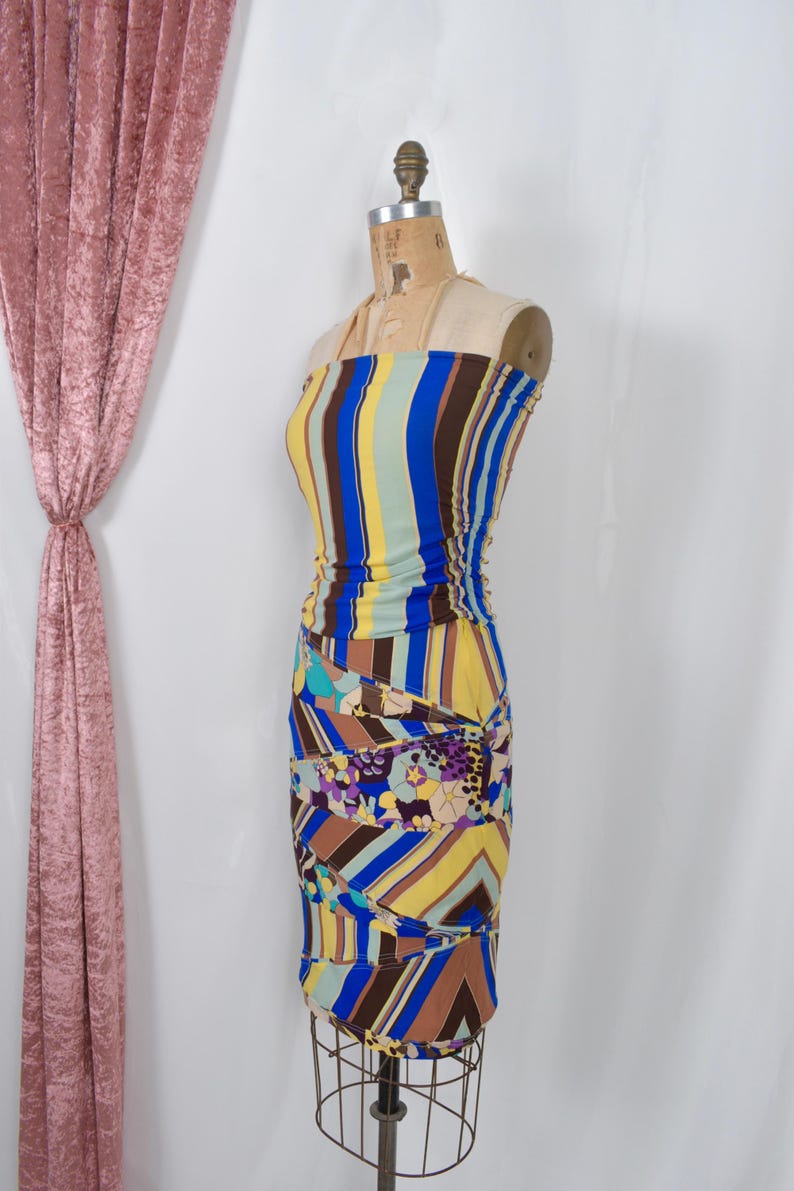 Puede incluir: Un vestido sin tirantes colorido con rayas verticales en azul, amarillo y marr&oacute;n. La parte inferior presenta un patr&oacute;n geom&eacute;trico y floral. El vestido se exhibe en un maniqu&iacute;, mostrando su dise&ntilde;o ajustado.
