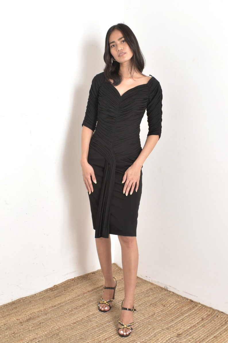 May include: A black, form-fitting dress with a sweetheart neckline and ruched detailing. The dress has a knee-length hem and three-quarter sleeves.