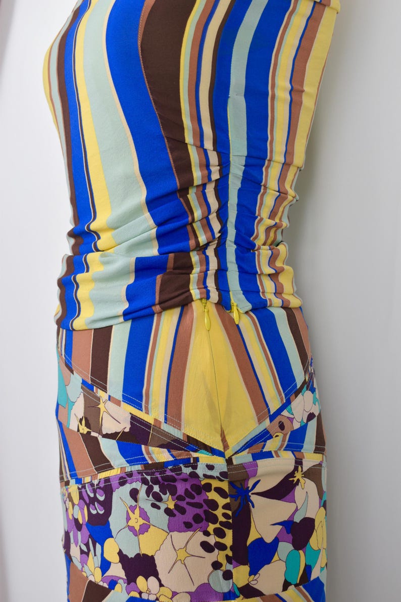 Puede incluir: Un vestido colorido y ajustado con rayas verticales azules, amarillas y marrones. La parte inferior presenta un estampado floral con detalles morados y beige. Una cremallera amarilla recorre el lateral.