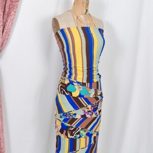 Puede incluir: Un vestido ajustado sin tirantes con rayas verticales amarillas, azules, marrones y verdes. La parte inferior presenta un estampado floral y abstracto. El vestido se exhibe en un maniqu&iacute; vintage.
