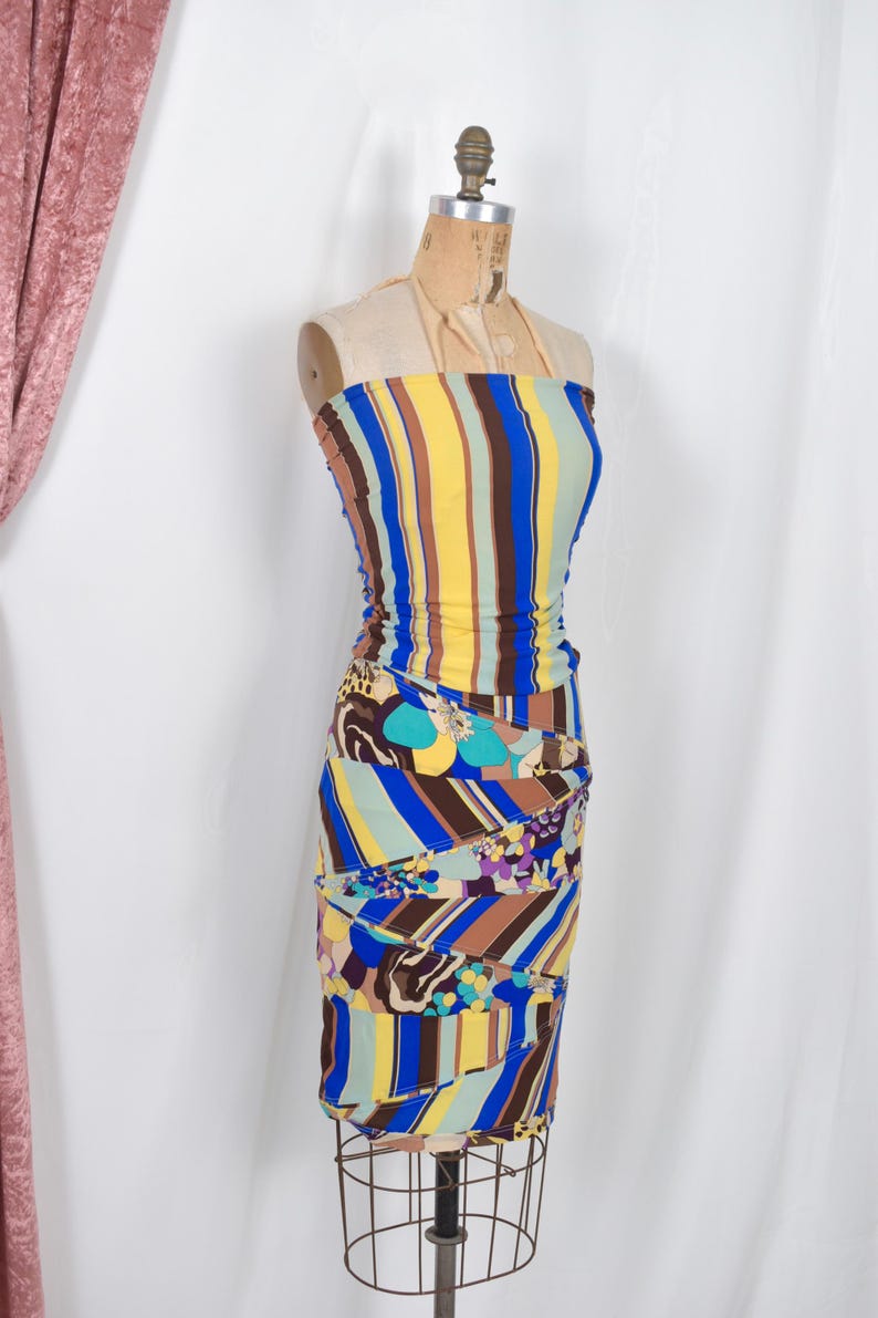 Puede incluir: Un vestido ajustado sin tirantes con rayas verticales amarillas, azules, marrones y verdes. La parte inferior presenta un estampado floral y abstracto. El vestido se exhibe en un maniqu&iacute; vintage.