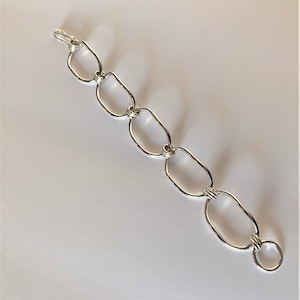 May include: A silver-toned bracelet featuring a series of interconnected, elongated oval links. The links are smooth and polished, creating a reflective surface. The bracelet has a simple hook clasp for closure, and is displayed on a white background.