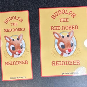 May include: Two yellow and red rectangular stickers with a cartoon image of Rudolph the Red-Nosed Reindeer. The text on the stickers reads "Rudolph the Red-Nosed Reindeer" and "@charmabudgets".