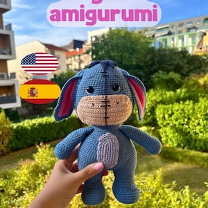 May include: A handmade blue Eeyore amigurumi plush toy with a tan muzzle and pink inner ears. The crochet figure has a purple tail and is held in front of a green background. The text "Eeyore amigurumi" is displayed above the toy.