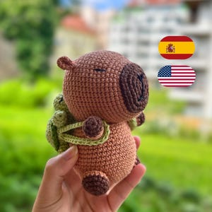 Könnte beinhalten: Ein gehäkeltes Capybara-Plüschtier in Brauntönen, mit einer dunkleren braunen Schnauze und Füßen. Es hat einen grünen Rucksack und wird in einer Hand gehalten. Das Plüschtier hat ein niedliches, freundliches Aussehen.