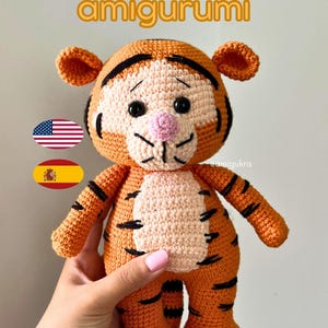 May include: A crocheted Tigger amigurumi toy in orange and black. The plush tiger has a light beige face and belly, black stripes, and a pink nose. The text "Tigger amigurumi" is displayed at the top of the image.