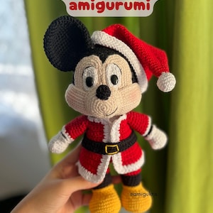 May include: A crocheted Mickey Mouse amigurumi wearing a Santa hat and suit. The figure has black ears, a cream-colored face, and is dressed in a red suit with white trim and a black belt. The text "Mickey Mouse amigurumi" is at the top.