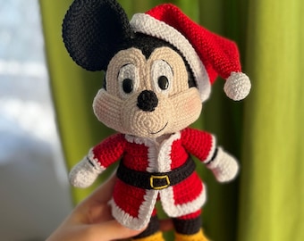 Może przedstawiać: Szydełkowe amigurumi Myszki Miki ubrane w strój Świętego Mikołaja. Postać ma czarne uszy, kremową twarz i nosi czerwony kapelusz i strój Świętego Mikołaja z białym wykończeniem i czarnym paskiem. Tekst "Mickey Mouse amigurumi" znajduje się na górze.