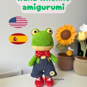 Könnte beinhalten: Handgefertigtes grünes Häkelfrosch-Amigurumi mit blauer Latzhose, roter Fliege und roten Schuhen. Der Text "Rana Rhenne amigurumi" steht über dem Frosch. Eine Sonnenblume und andere Dekorationen befinden sich im Hintergrund.