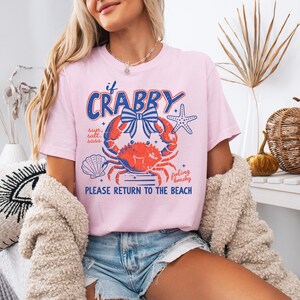 May include: A light pink t-shirt with a graphic design featuring a crab, starfish, and seashell. The text on the shirt reads "if Crabby" and "Please Return to the Beach". The crab is red and blue.