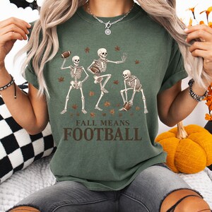 May include: Green t-shirt with a graphic of three skeleton figures playing football, surrounded by autumn leaves. The text "FALL MEANS FOOTBALL" is printed below the skeletons. The shirt is a casual, short-sleeved style.