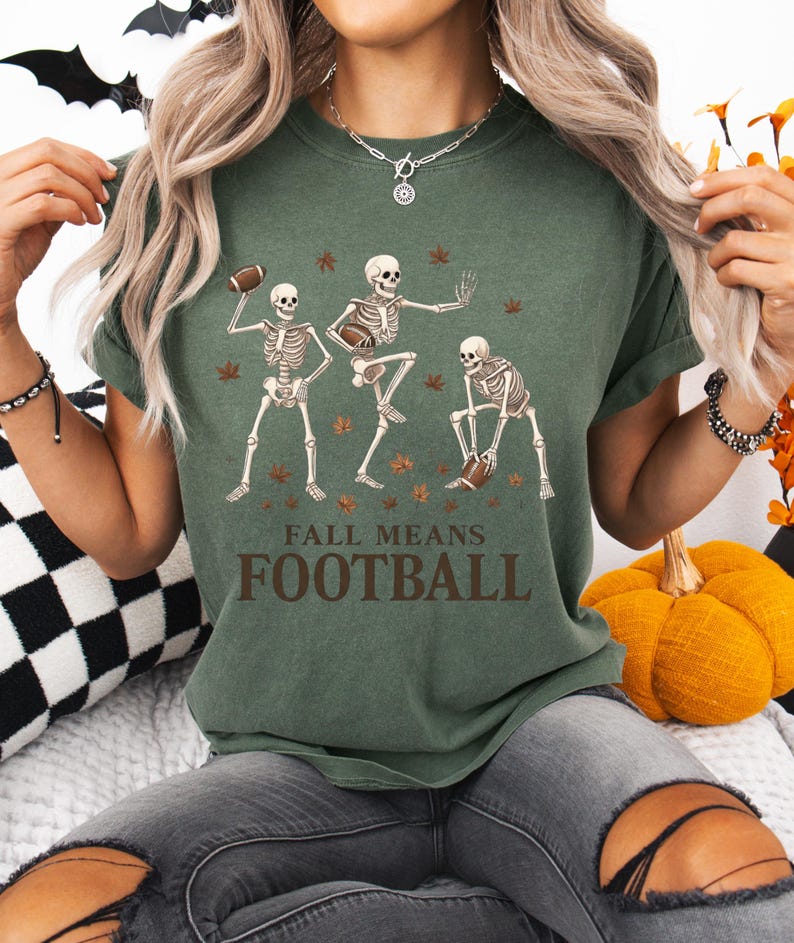 May include: Green t-shirt with a graphic of three skeleton figures playing football, surrounded by autumn leaves. The text "FALL MEANS FOOTBALL" is printed below the skeletons. The shirt is a casual, short-sleeved style.