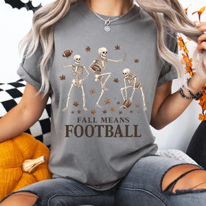 May include: Gray t-shirt with a graphic of three skeleton figures playing football, surrounded by autumn leaves. The text "FALL MEANS FOOTBALL" is printed below the skeletons. The shirt is a casual, fall-themed design.