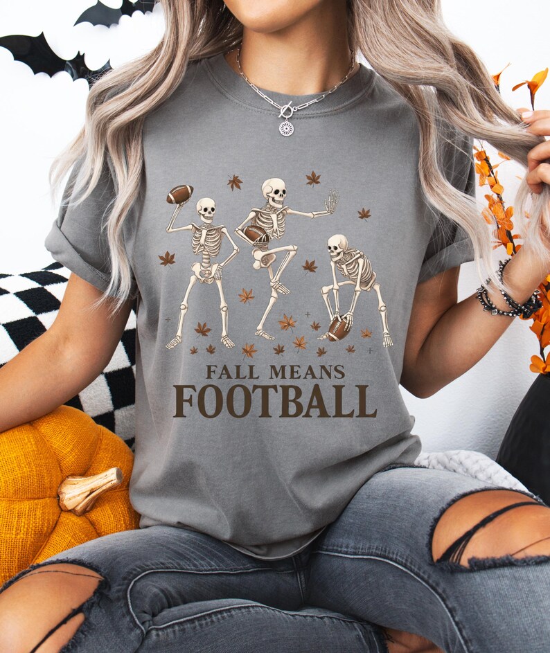 May include: Gray t-shirt with a graphic of three skeleton figures playing football, surrounded by autumn leaves. The text "FALL MEANS FOOTBALL" is printed below the skeletons. The shirt is a casual, fall-themed design.