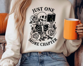Pode incluir: Camisola creme com um desenho preto de um esqueleto lendo um livro, rodeado por rosas. O texto diz "JUST ONE MORE CHAPTER". Uma caneca laranja é segurada na mão.