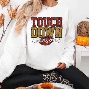 May include: White sweatshirt with the text "TOUCH DOWN season" in red and gold glitter lettering, with a football graphic. The sweatshirt is a crew neck style.