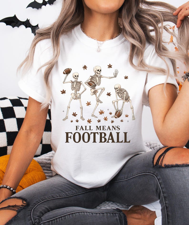 May include: White t-shirt featuring a graphic of three skeleton figures playing football, with brown autumn leaves. The text "FALL MEANS FOOTBALL" is printed below the skeletons. A fun, seasonal design.
