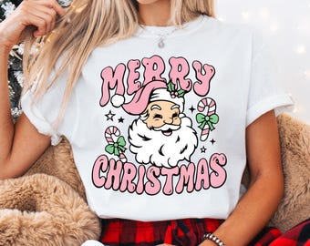 May include: White t-shirt with a retro "Merry Christmas" graphic in pink. The design features Santa Claus with a pink hat, candy canes, and stars. A festive holiday tee.