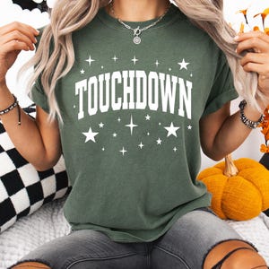 以下が含まれることがあります： 白い星に囲まれた、白いブロック体の「TOUCHDOWN」という文字が入った濃い緑色のTシャツ。 このシャツは半袖のクルーネックです。 モデルはグレーのジーンズを着用しています。