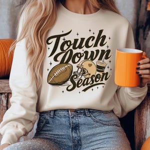 May include: Cream-colored sweatshirt with the text "Touch Down Season" and football-themed graphics. The design includes a football, helmet, and a drink cup. The person in the image is holding an orange mug.