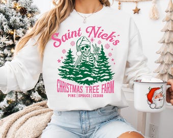 May include: White sweatshirt with a graphic that reads "Saint Nick's Christmas Tree Farm" in pink and green. The graphic features a skeleton wearing a Santa hat and holding skis. The sweatshirt is paired with blue jeans.