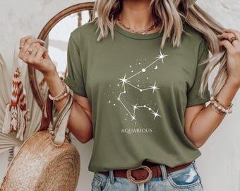 Könnte beinhalten: Olivgrünes T-Shirt mit weißem Sternbild-Design und dem Wort "AQUARIOUS" darunter. Das Shirt hat kurze Ärmel und einen Rundhalsausschnitt. Das Model trägt eine blaue Jeans und einen braunen Gürtel.