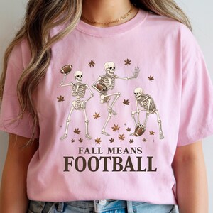 May include: A light pink t-shirt featuring a graphic of three skeleton figures playing football, surrounded by autumn leaves. The text "FALL MEANS FOOTBALL" is printed in a bold, dark brown font below the skeletons.