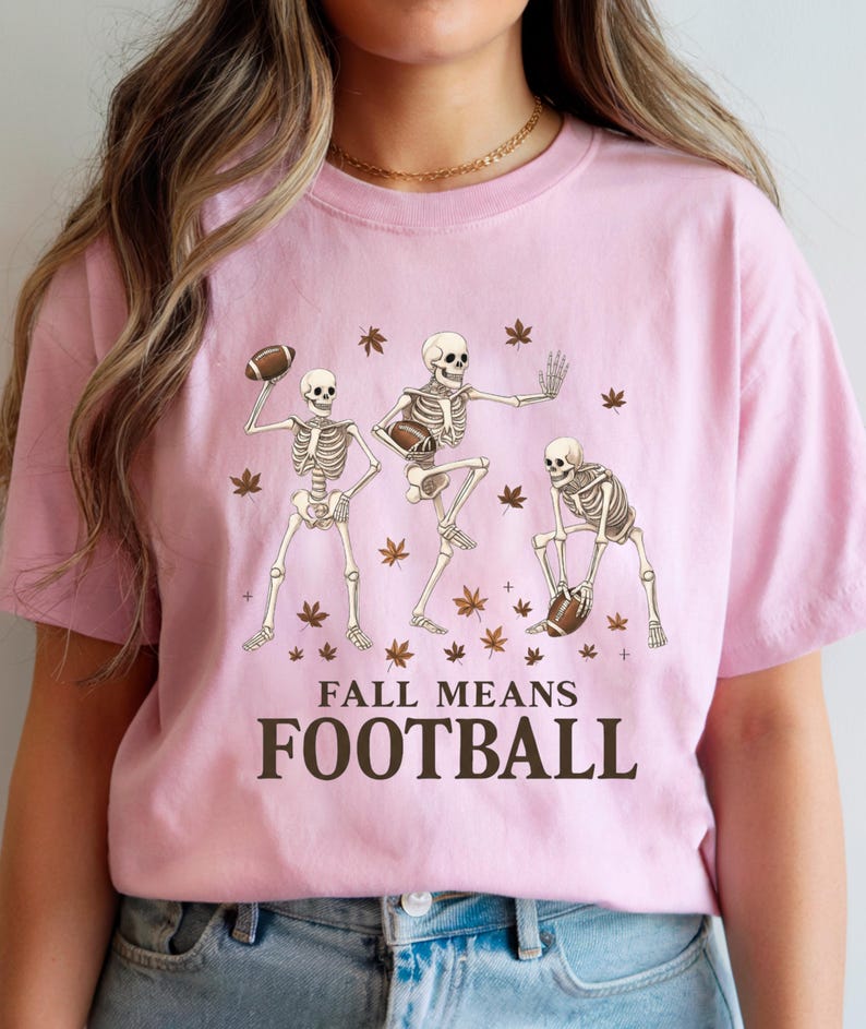 May include: A light pink t-shirt featuring a graphic of three skeleton figures playing football, surrounded by autumn leaves. The text "FALL MEANS FOOTBALL" is printed in a bold, dark brown font below the skeletons.