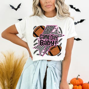 May include: White t-shirt with a graphic design featuring two brown footballs and a lightning bolt with a leopard print and checkered pattern. The text "Game Day Baby!" is in pink and white.