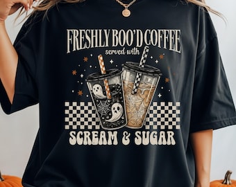 May include: Black t-shirt with a Halloween-themed graphic. The design features two iced coffee drinks with straws, one with a ghost and spiderweb, the other with a checkered pattern. Text reads "FRESHLY BOO'D COFFEE served with SCREAM & SUGAR."
